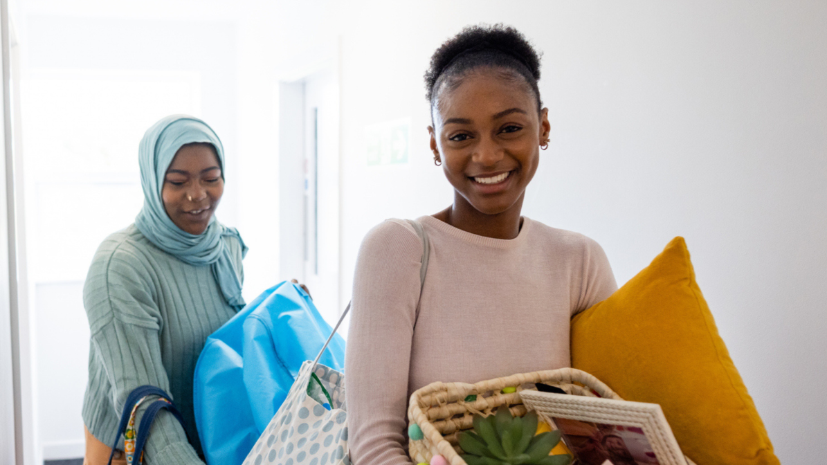 Two students walk through a hallway, each carrying boxes full of random items, as they move into their new accommodation.