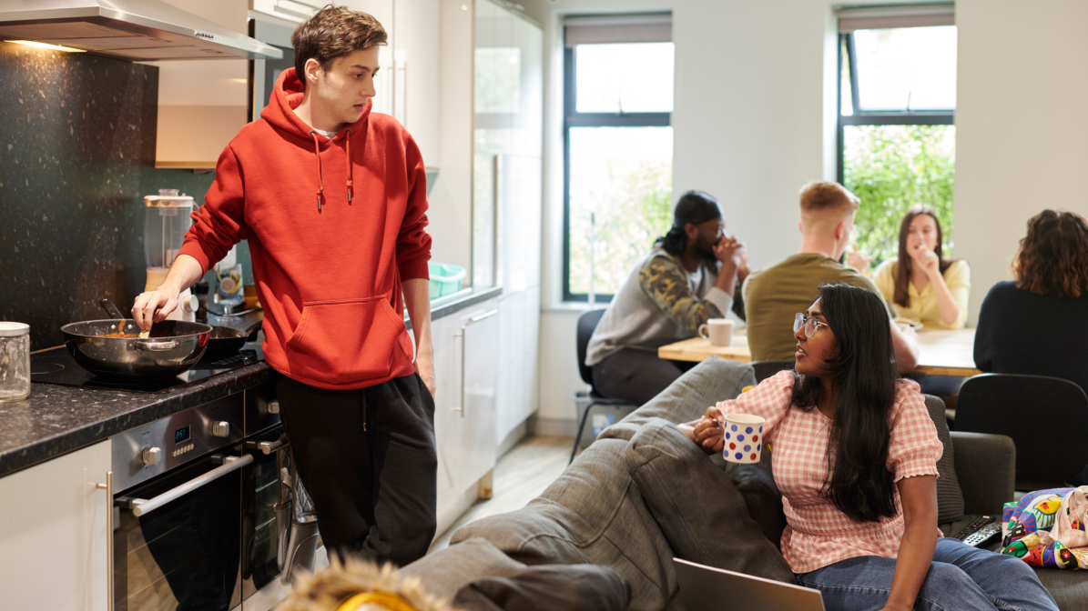 A group of students in a living room, some engaged with a laptop, others cooking, showcasing a casual study space.