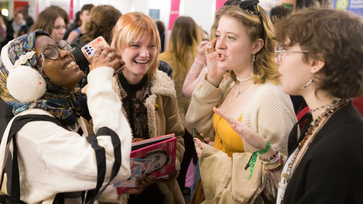 The image shows four young people smiling and chatting at a UCAS event. One is holding a phone and another a book. They are gesturing and engaged in conversation.