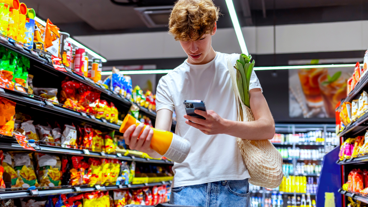 A male student wearing a white t-shirt and blue just standing in the aisle of a supermarket holding a bottle of orange juice whilst looking at his mobile phone 