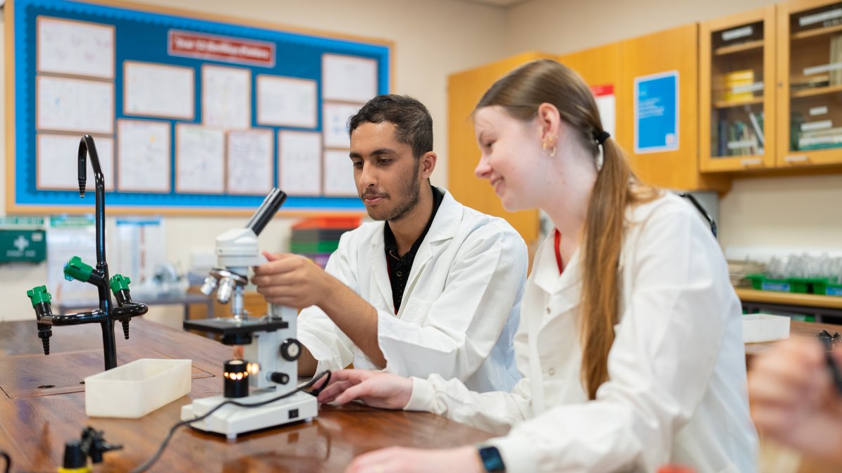 Two students wearing lab coats sat in a classroom using a microscope