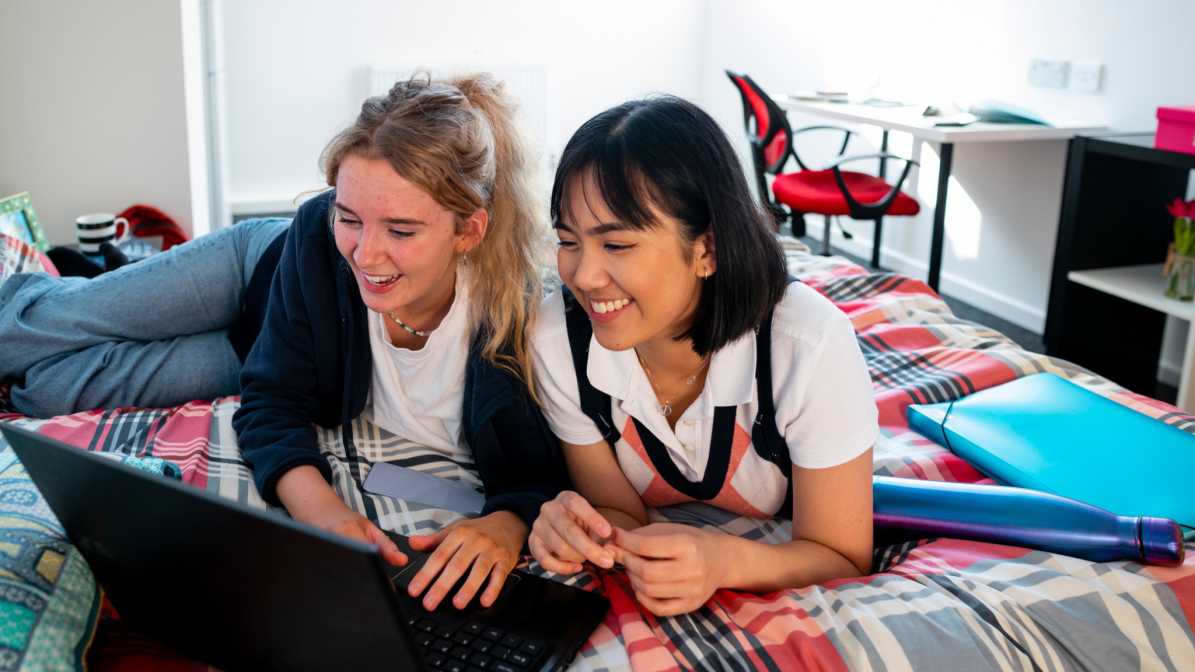 Two students laid on a bed, looking at a laptop