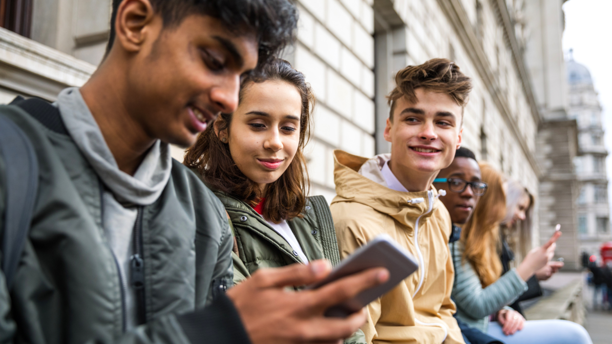 Five students socialising on a city bench, one using a smartphone — a real-world glimpse into how next-gen consumers connect and engage, offering brands insight into reaching student audiences through mobile and social platforms.
