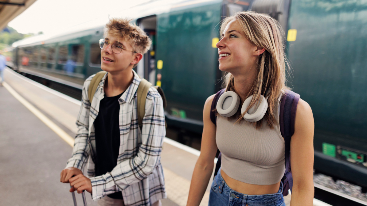 Two gap year students with backpacks and travel gear smiling on a train platform, representing UK 18-year-olds preparing for travel, volunteering, or career experiences. Ideal visual for brands targeting students during their year out through campaigns in education, travel, and youth marketing sectors.
