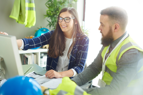 Two construction workers, one a woman in a suit and the other a man in hi vis jacket, sat at a desk looking at a computer screen
