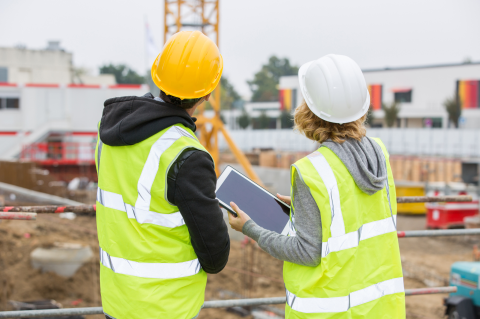 Two construction workers, wearing hi vis jackets and helmets, stood outside looking at a construction site