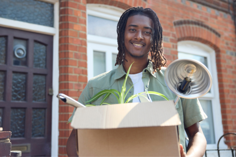 A student stood outside the front door of a terraced house, holding a box containing a plant and lamp